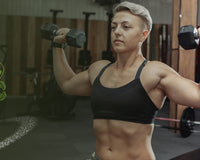 Woman performing a standing dumbbell shoulder press in a gym with weight racks and a barbell in the background