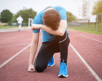 Runner kneeling on an outdoor track holding his face in apparent exhaustion with another runner in the background