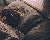 Person sleeping on their side under a knitted blanket in a dimly lit bedroom near closed blinds