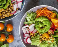 Bowl of raw vegetable salad with sliced avocado, chickpeas, radish, carrot ribbons and cherry tomatoes on a gray stone surface